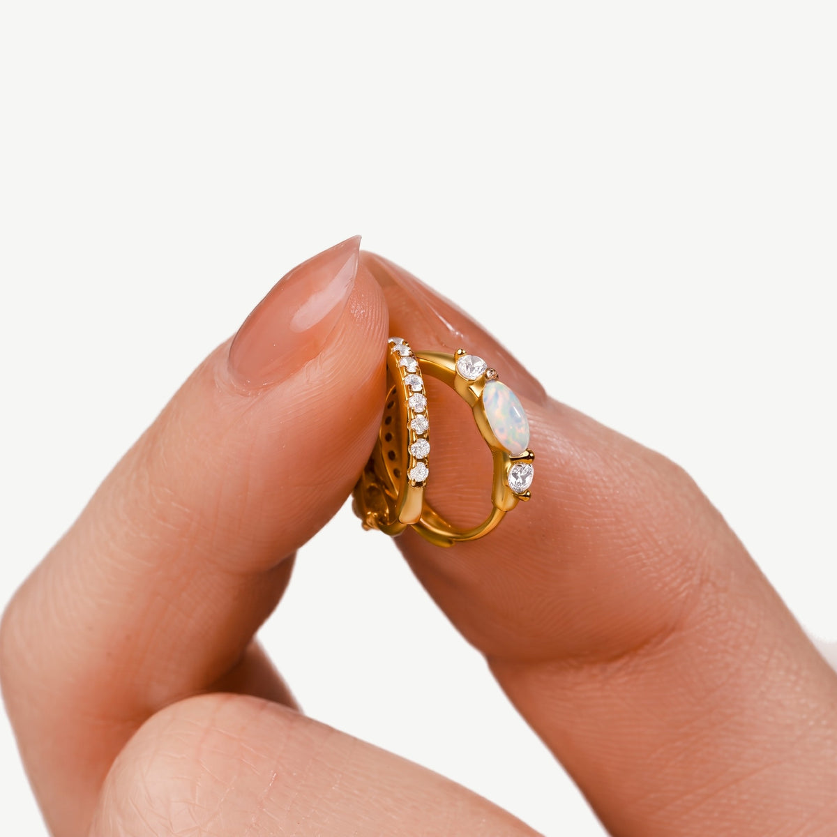 Fingers holding a pair of gold plated opal huggie hoop earrings up close, marquise opal and cubic zirconia band