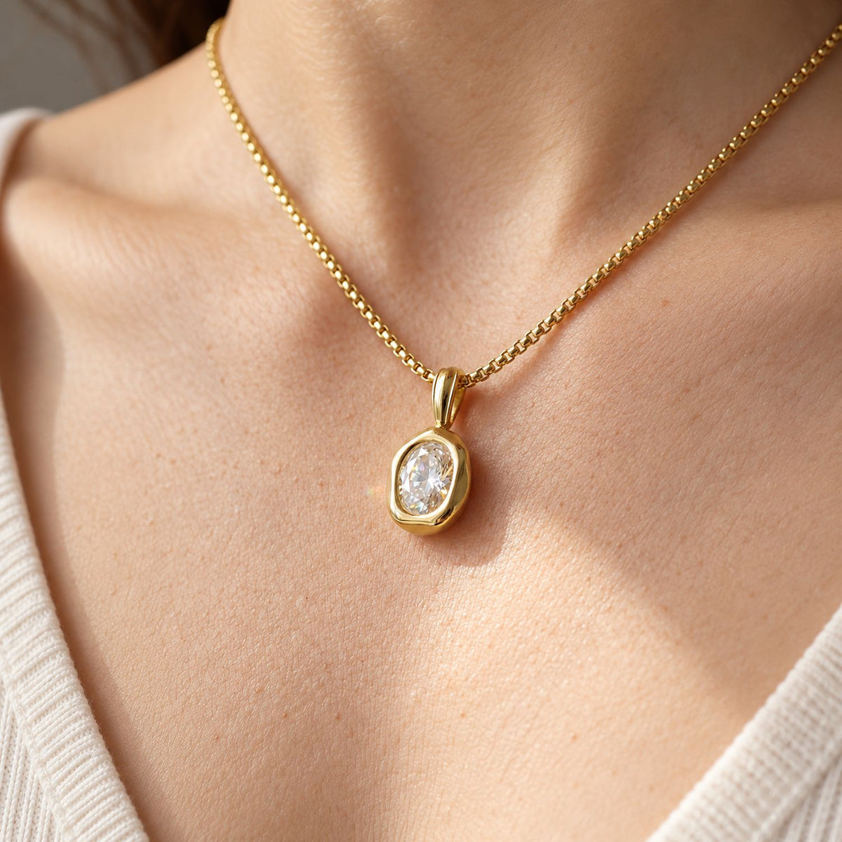 Close view of a 14k gold plated oval pendant necklace on a box chain at the collarbone, worn with an ivory knit top