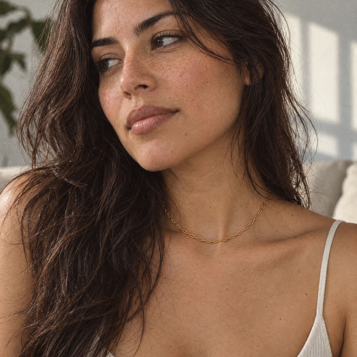 Woman with long dark wavy hair in an ivory tank wearing a dainty gold paperclip chain necklace at the base of her neck