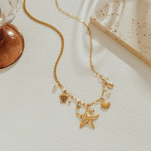 Gold plated seashell charm necklace laid on white stone with a hammered starfish, sea turtle, shells and pearls