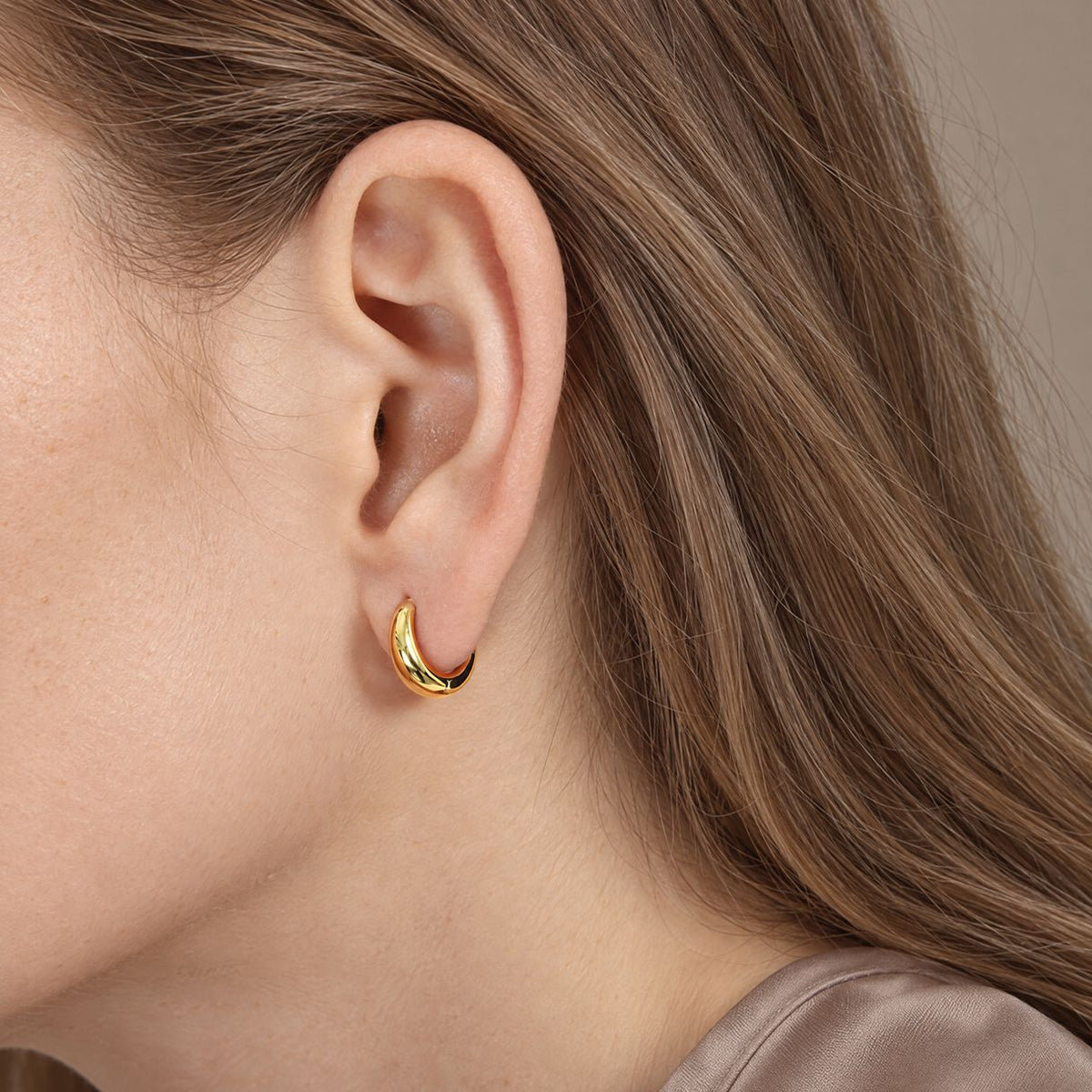 Side view of a 10 mm gold-plated teardrop huggie hoop worn on the earlobe, light-brown hair and taupe silk top