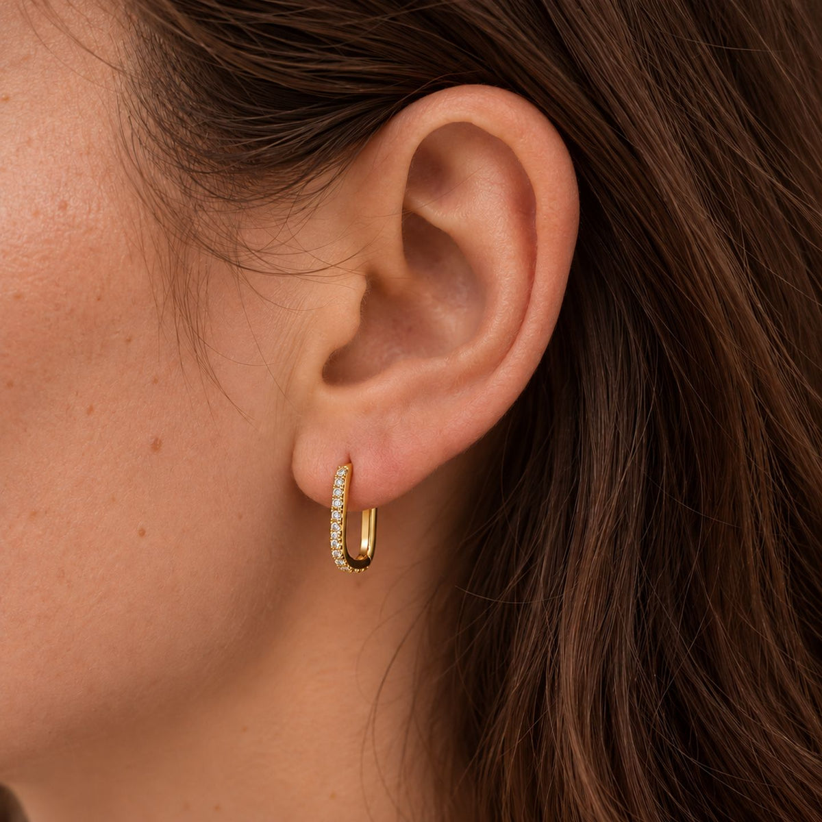 Close crop of a woman's ear and cheek, long brown hair, with a gold plated pavé U hoop huggie worn in the lobe