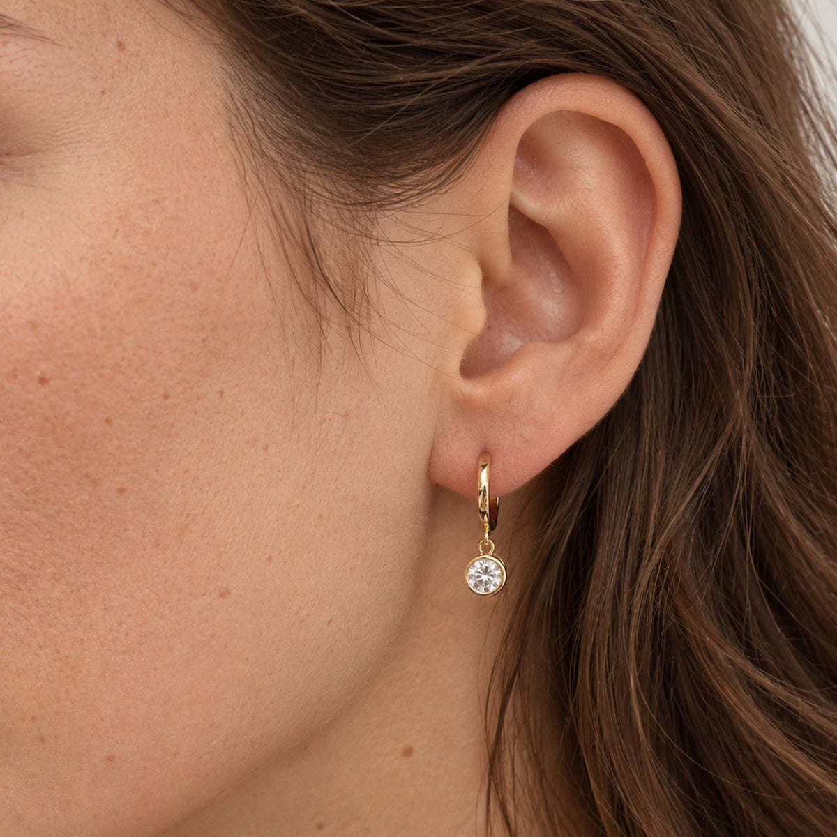 Close view of a woman's ear and jaw, a 0.5 ct moissanite drop hanging from a small 14k gold plated huggie hoop