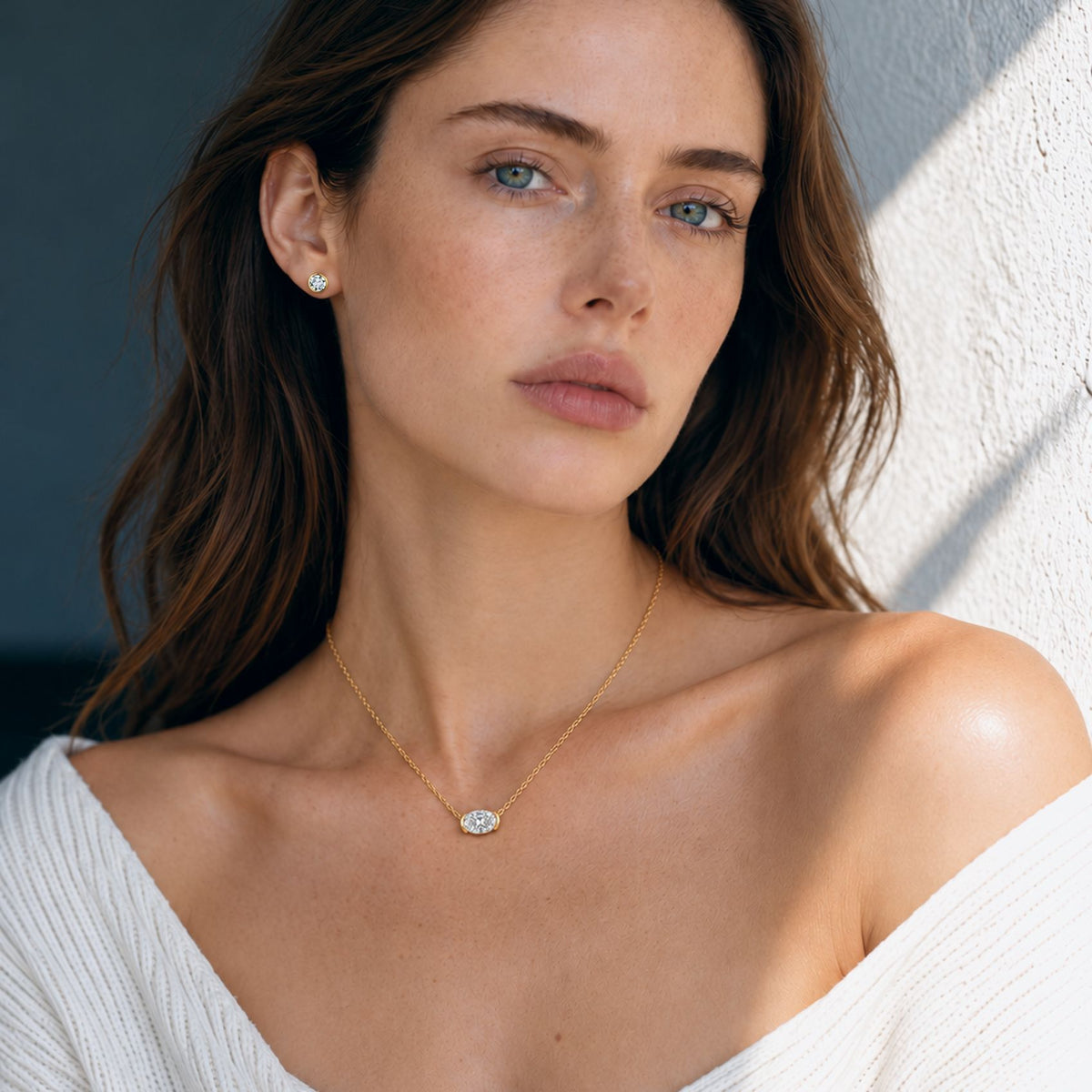 Woman wearing a 0.5 ct gold bezel moissanite stud earring with an oval pendant, sunlit portrait by a white wall
