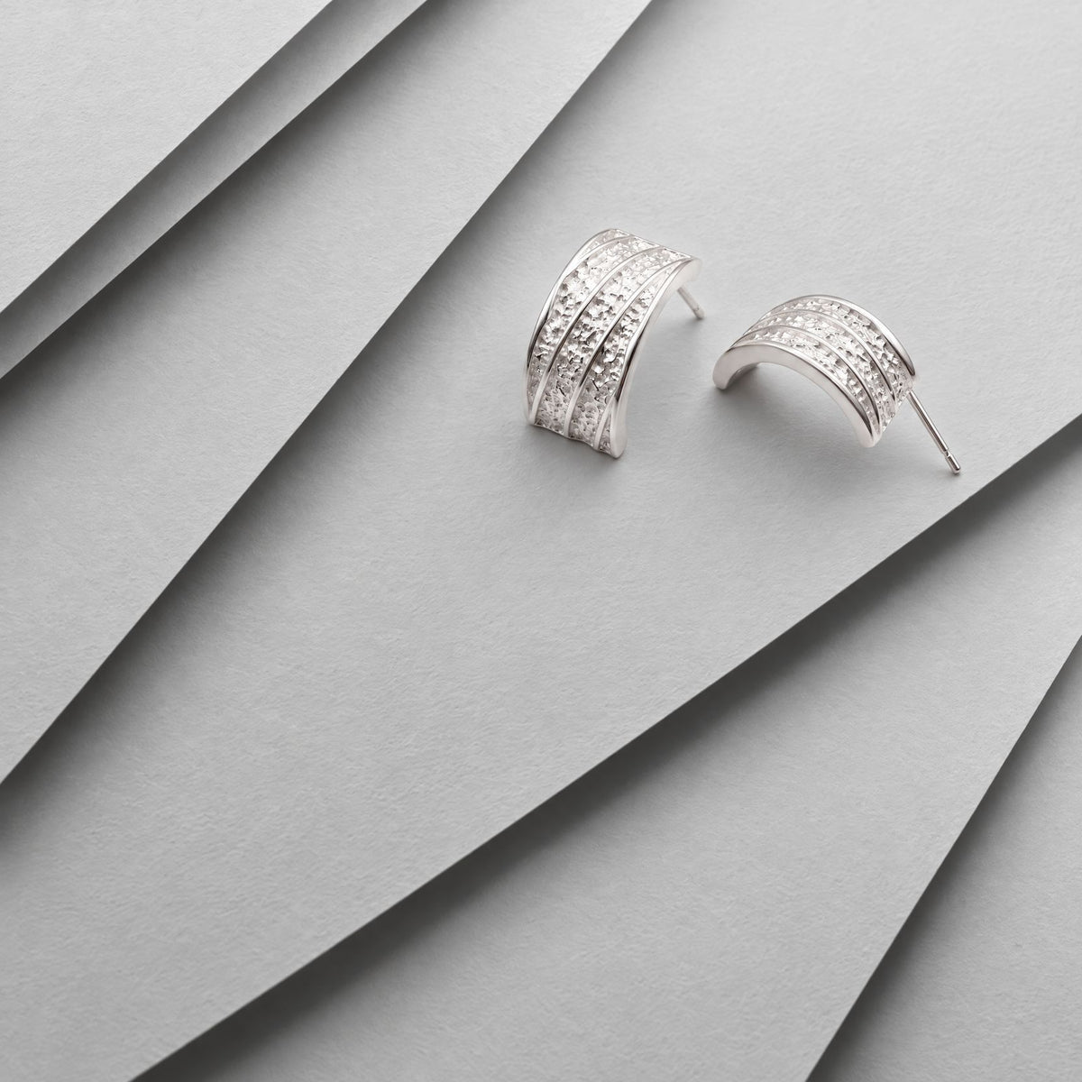 Chunky sterling silver hammered half hoop earrings on layered grey card, one showing the ribbed front, one the arc