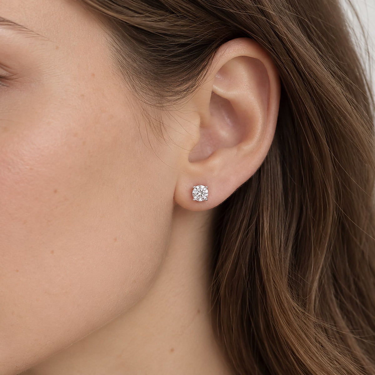 Close crop of a woman's ear and cheek, long brown hair, a round moissanite stud in sterling silver worn in her lobe