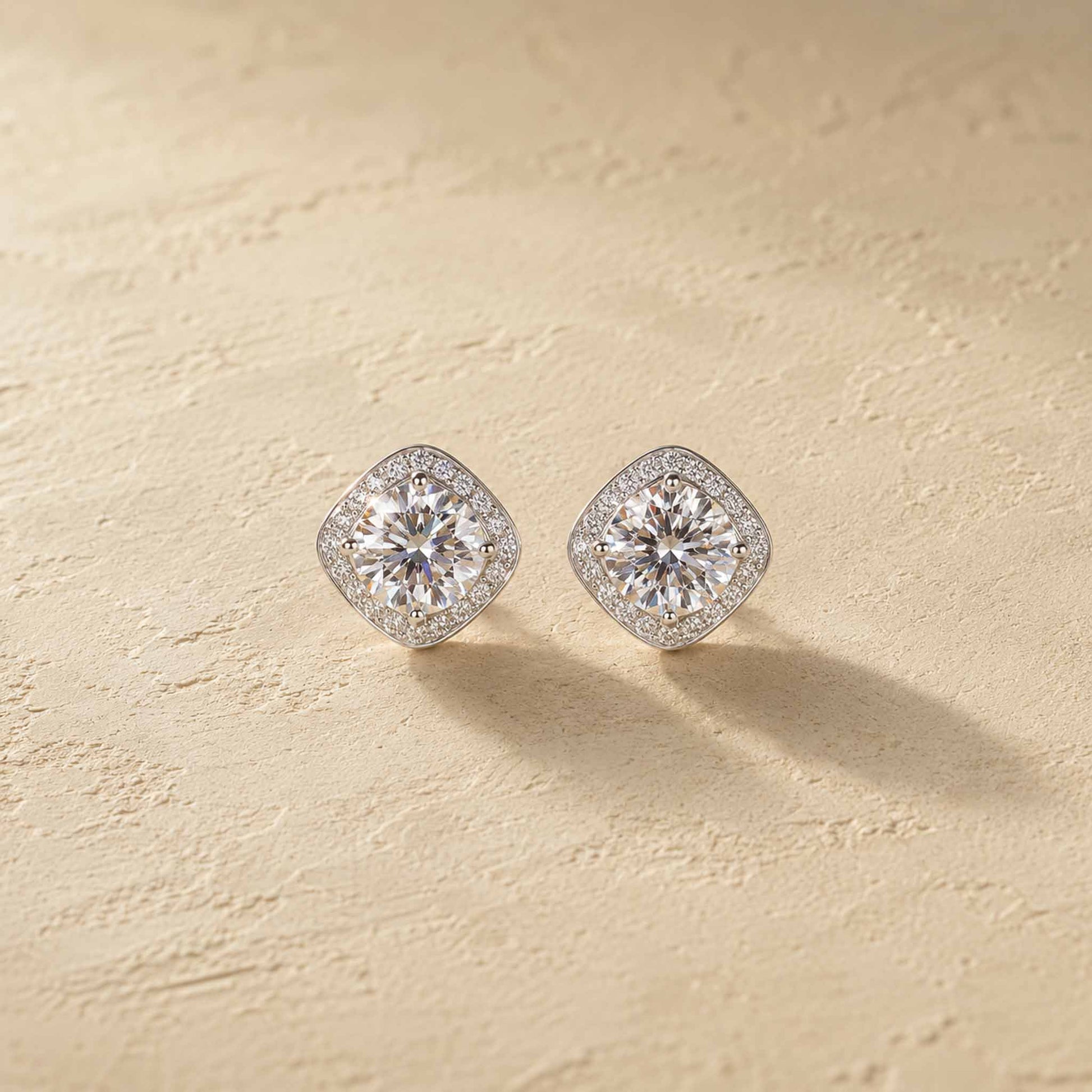 Pair of 1ct white moissanite halo stud earrings in sterling silver, cushion-shaped halos on warm cream surface