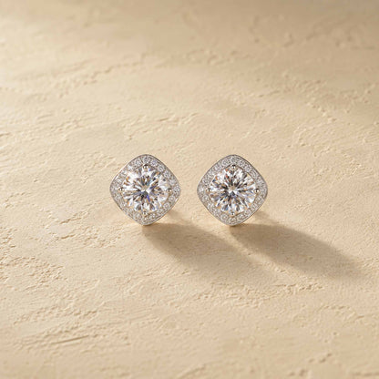 Pair of 1ct white moissanite halo stud earrings in sterling silver, cushion-shaped halos on warm cream surface