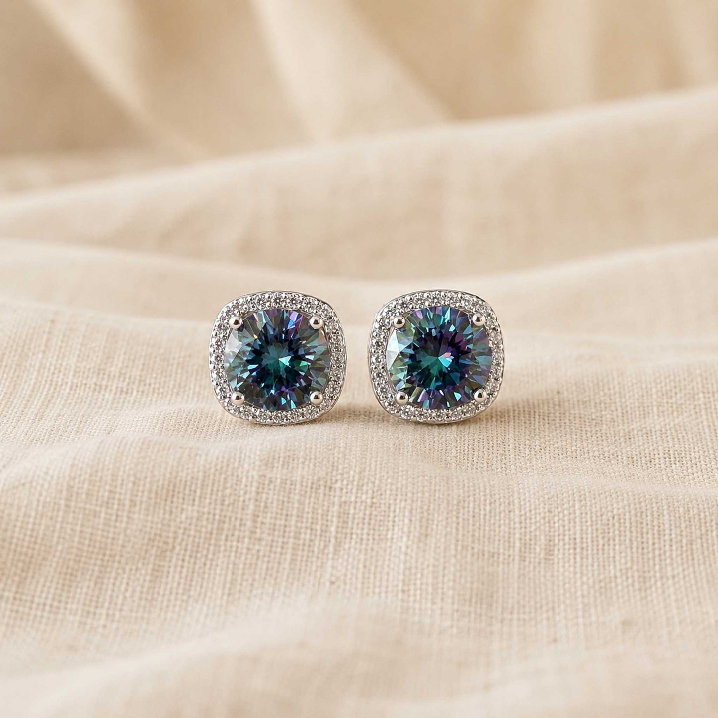 Pair of 1ct round cut rainbow blue moissanite halo stud earrings in sterling silver on cream linen