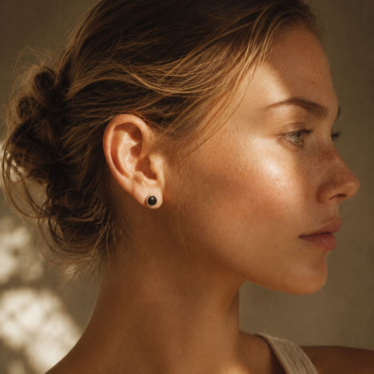 Woman wearing sterling silver black onyx stud earring with Greek key engraved bezel, shown in warm natural light with soft dappled shadows.