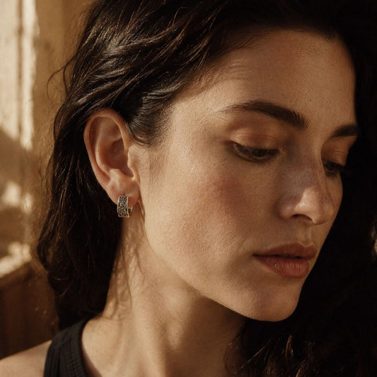 Female model wearing Museori sterling silver gothic cross and fleur-de-lis huggie hoop earring in warm afternoon light.