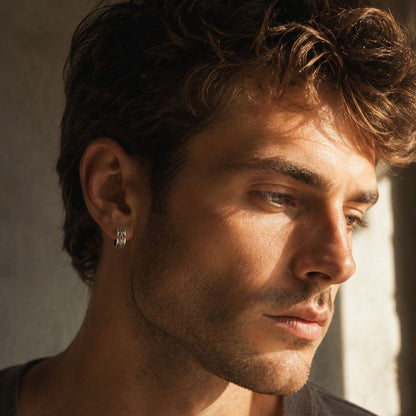 Male model wearing Museori sterling silver gothic cross and fleur-de-lis huggie hoop earring, warm sunlit profile.
