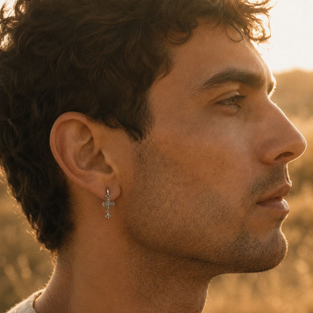 Sterling silver huggie hoop with a Gothic cross drop earring, worn by a man shown in profile at golden hour. Dark curly hair, light stubble, warm sunlit field behind. Soft editorial menswear mood.