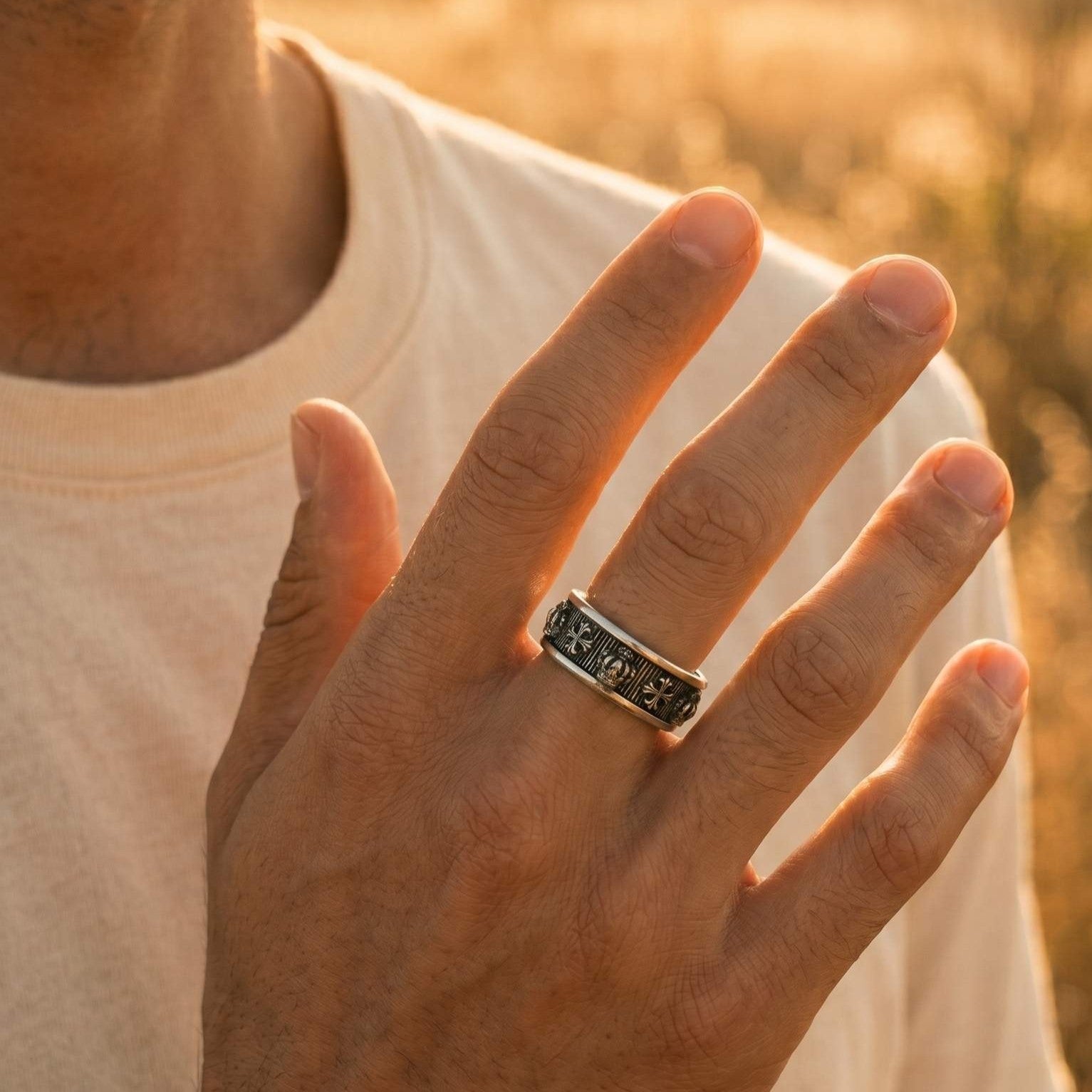 A man's hand raised near his chest wearing a men's antiqued sterling silver gothic ring with an alternating crown and cross engraving on an adjustable band, in warm golden-hour light.