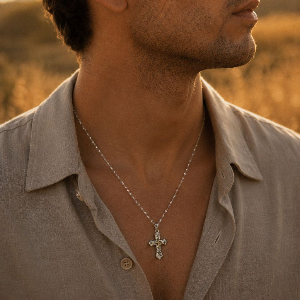 Man in an open-collar shirt wearing a men's oxidized sterling silver cross pendant necklace with a raised gold-tone center on a fine silver chain, resting on his chest in warm golden-hour light.