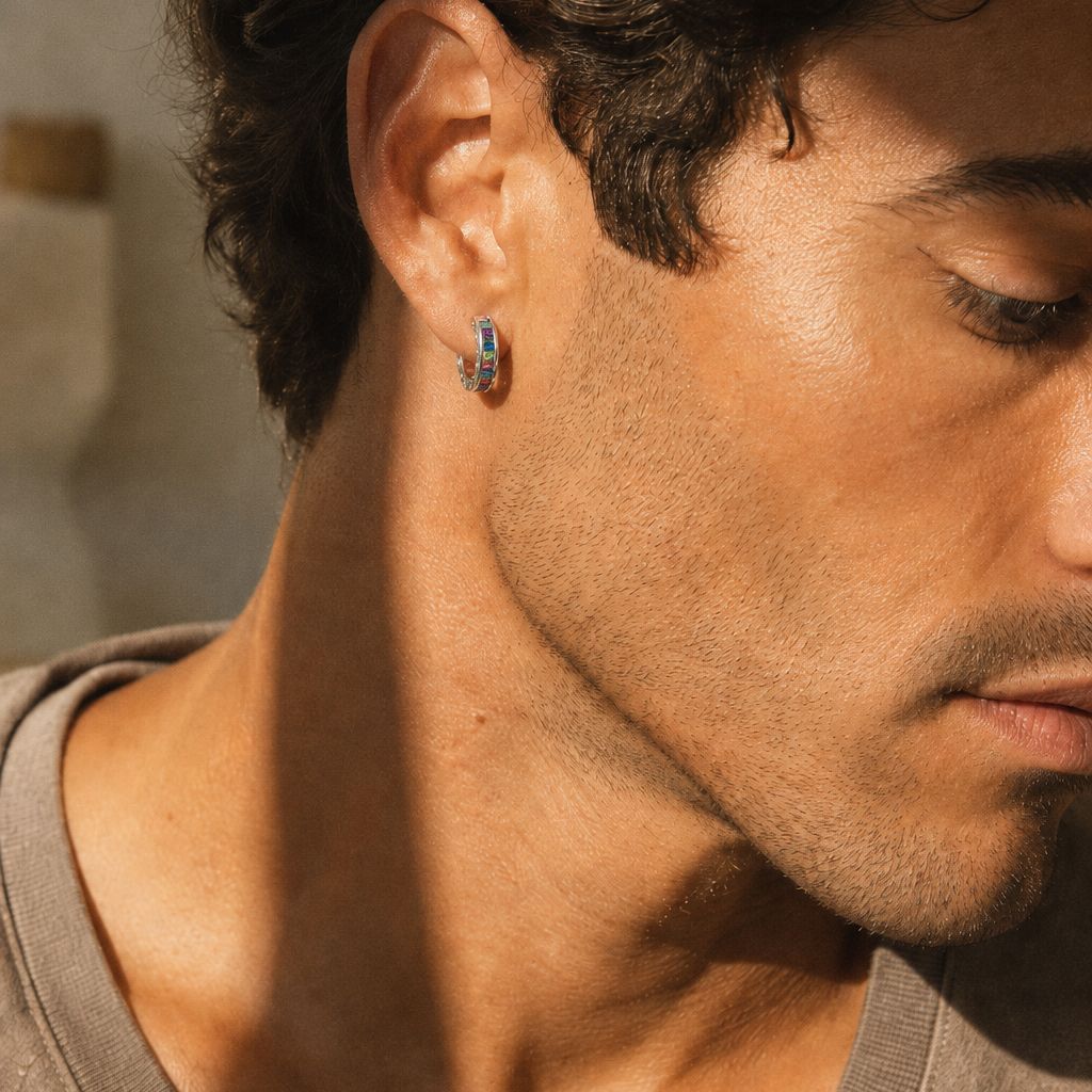 Man wearing sterling silver rainbow baguette huggie earring with multicolor stones and oxidized floral border, shown in warm natural light against a soft neutral background.