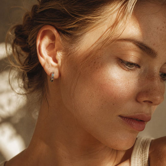 Woman wearing sterling silver rainbow baguette huggie earring with multicolor stones and oxidized floral border, shown in warm natural light with soft dappled shadows.