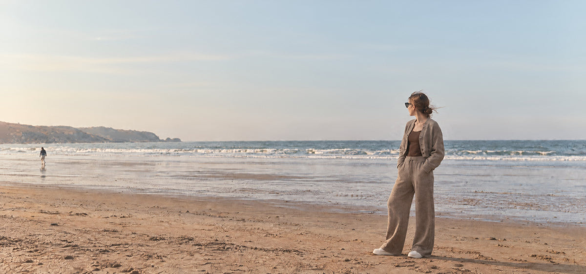 Woman in a taupe knit set and sunglasses standing on a wide beach at low tide under a pale sky