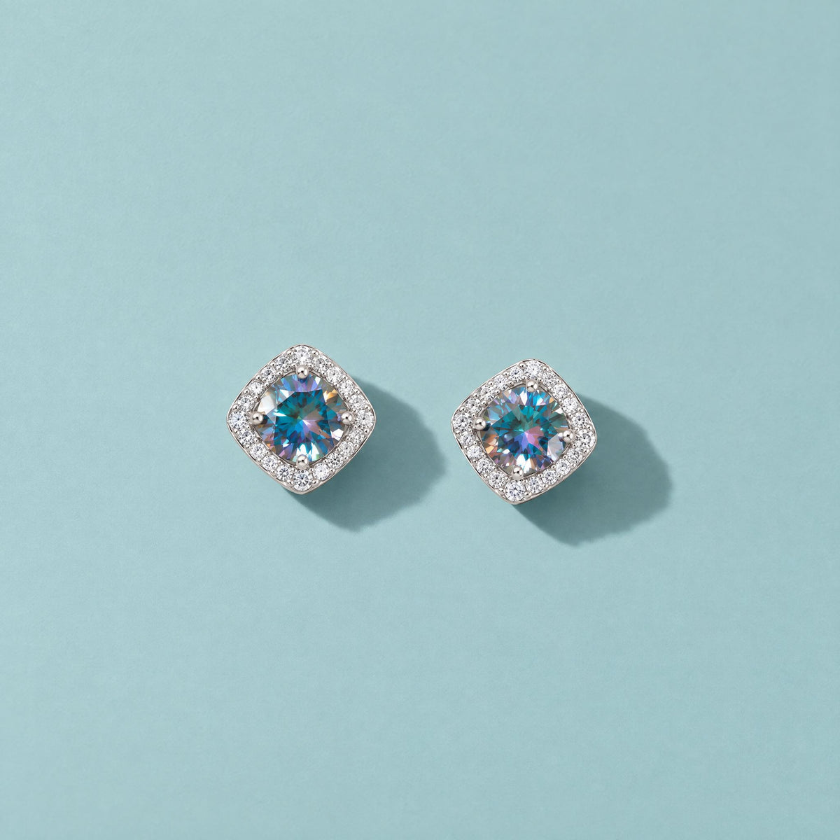 Rainbow blue moissanite halo stud earrings in sterling silver lying face up on a pale teal mist backdrop