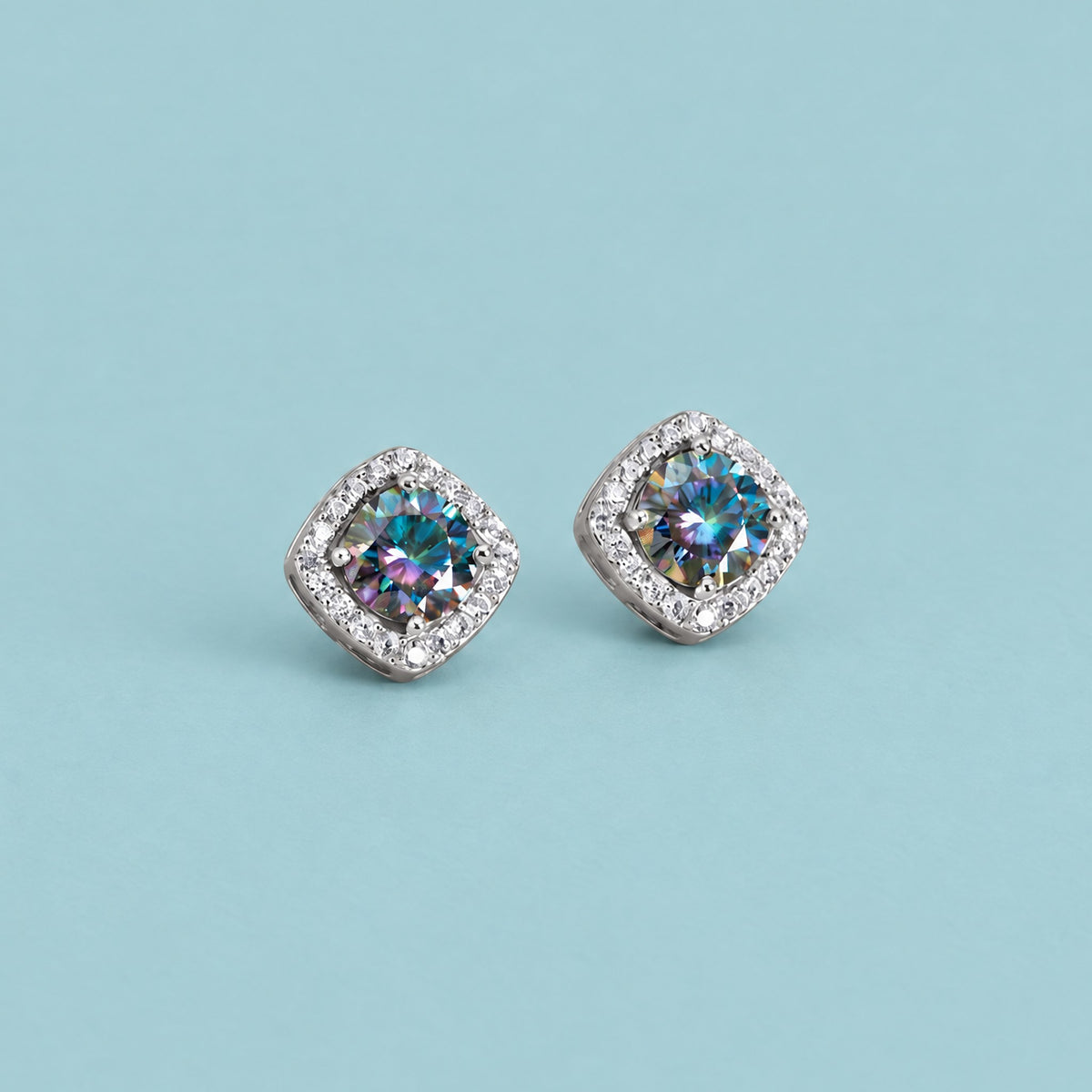 Rainbow blue moissanite halo stud earrings in sterling silver lying flat on a soft teal backdrop, cushion halo