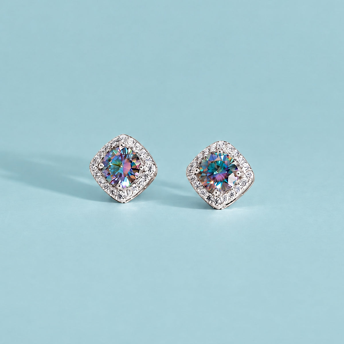 Pair of rainbow blue moissanite halo studs facing the camera on a soft teal backdrop, color shifting center stones