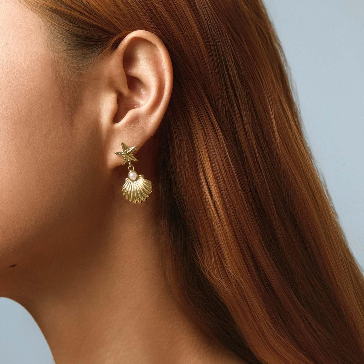 Woman with long auburn hair wearing a gold starfish and seashell drop earring on her lobe, side profile