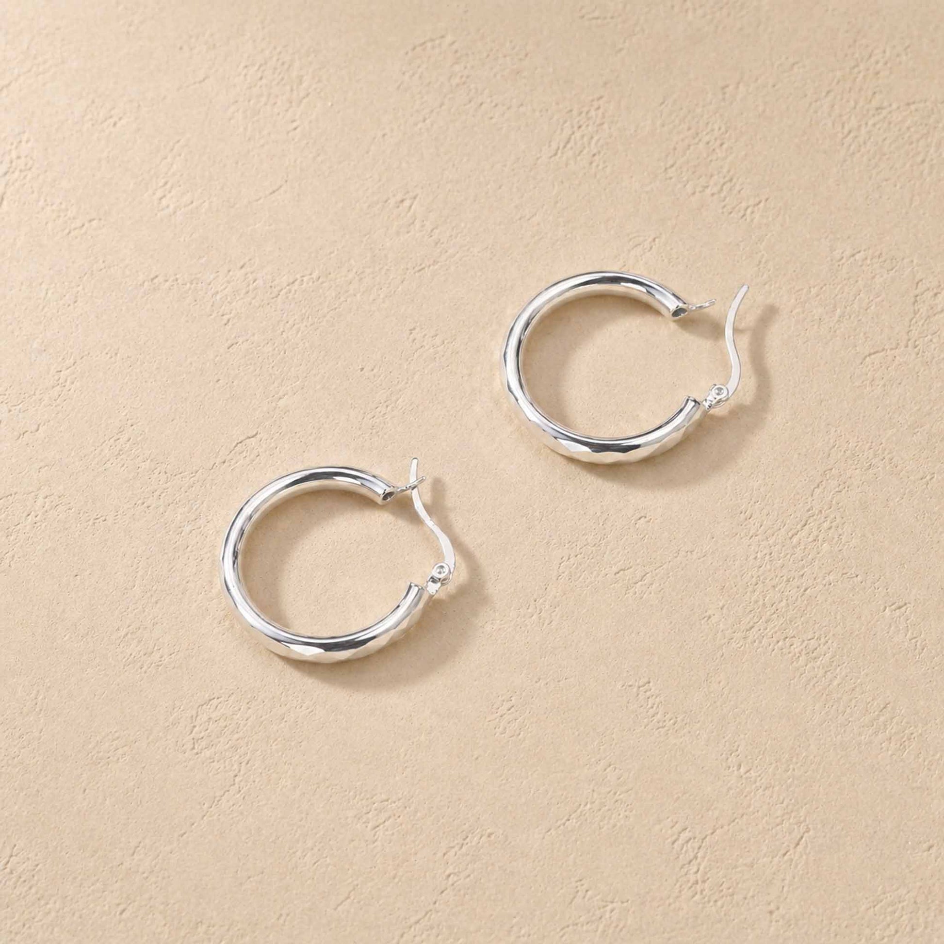 Faceted sterling silver hoop earrings shown open, top-down on warm cream background