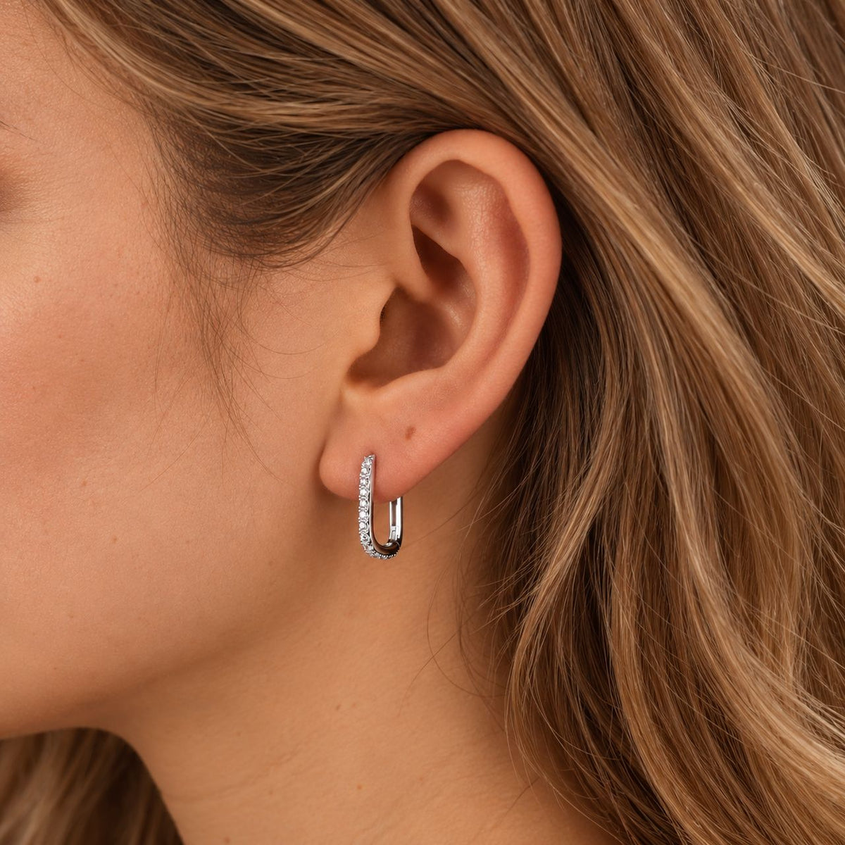Side view of a woman's ear and jaw with a sterling silver pavé huggie hoop in the lobe, honey blonde hair behind