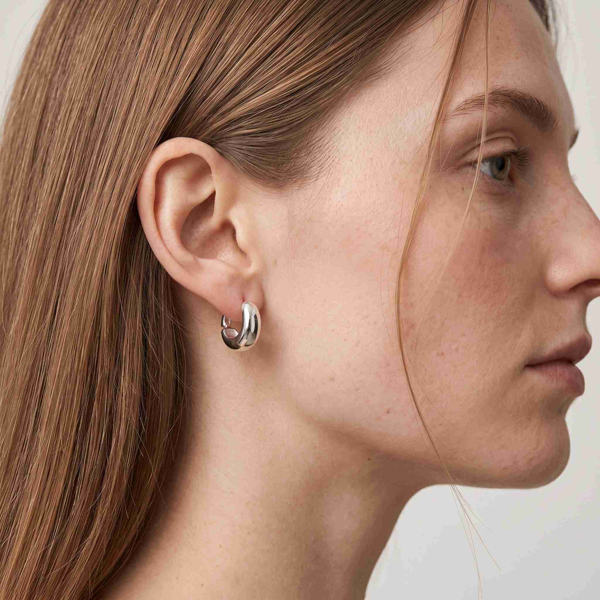 Woman wearing a sterling silver chunky dome huggie hoop earring, side profile view on model