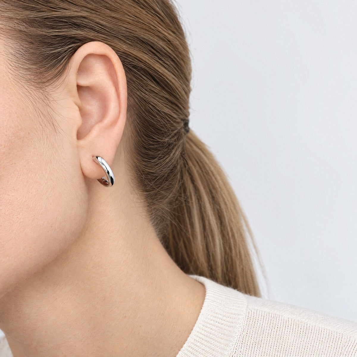Sterling silver tapered huggie hoop worn on the earlobe, side view of a model with slicked-back hair and ivory top