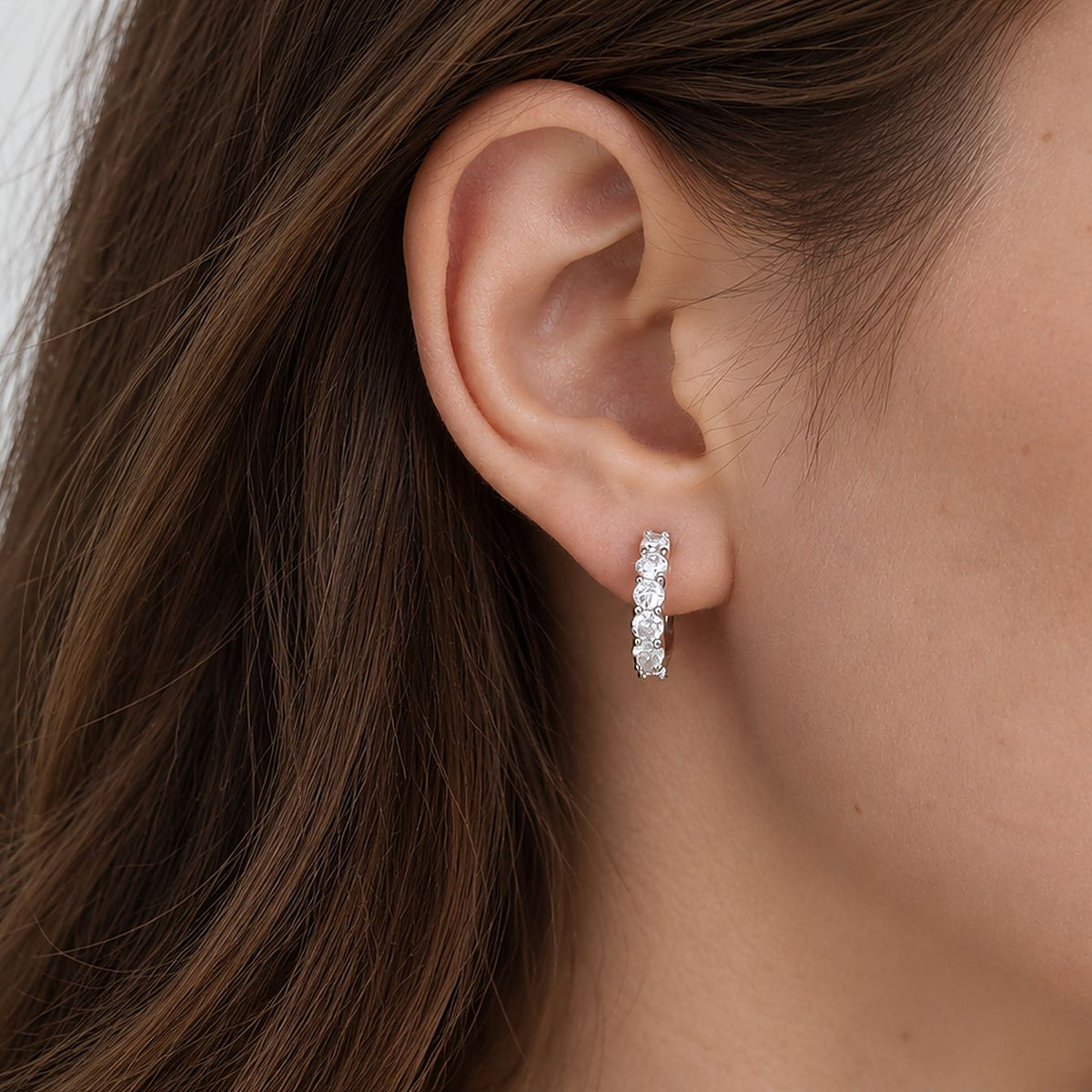 Woman with long brown hair wearing a sterling silver moissanite huggie hoop in her earlobe, close crop of ear and cheek