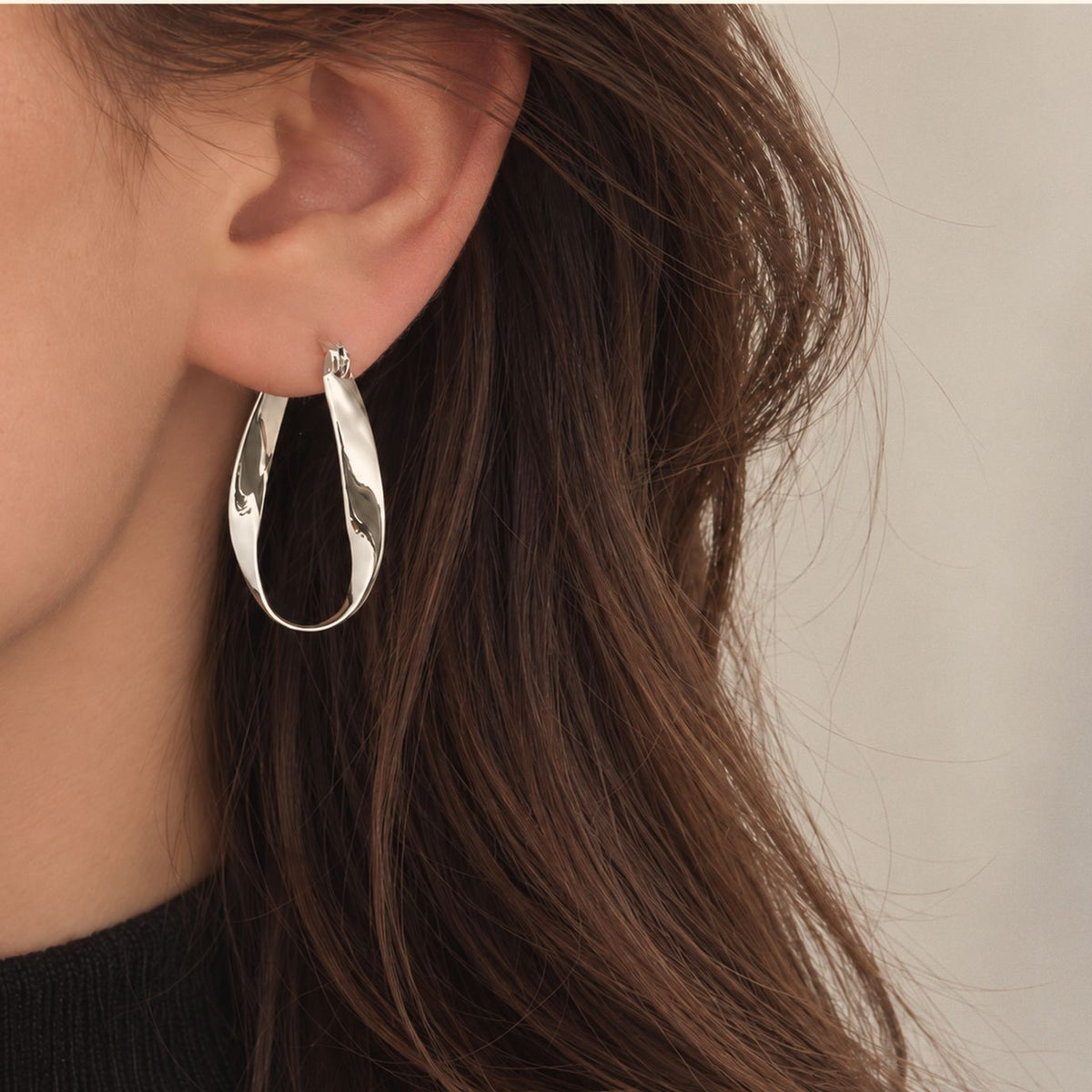 Close view of a twisted sterling silver hoop earring on the earlobe, polished teardrop band against brown hair