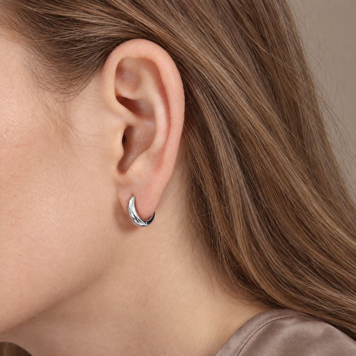 10 mm silver teardrop huggie hoop worn on the earlobe, side view of a model in a taupe silk top with hair down