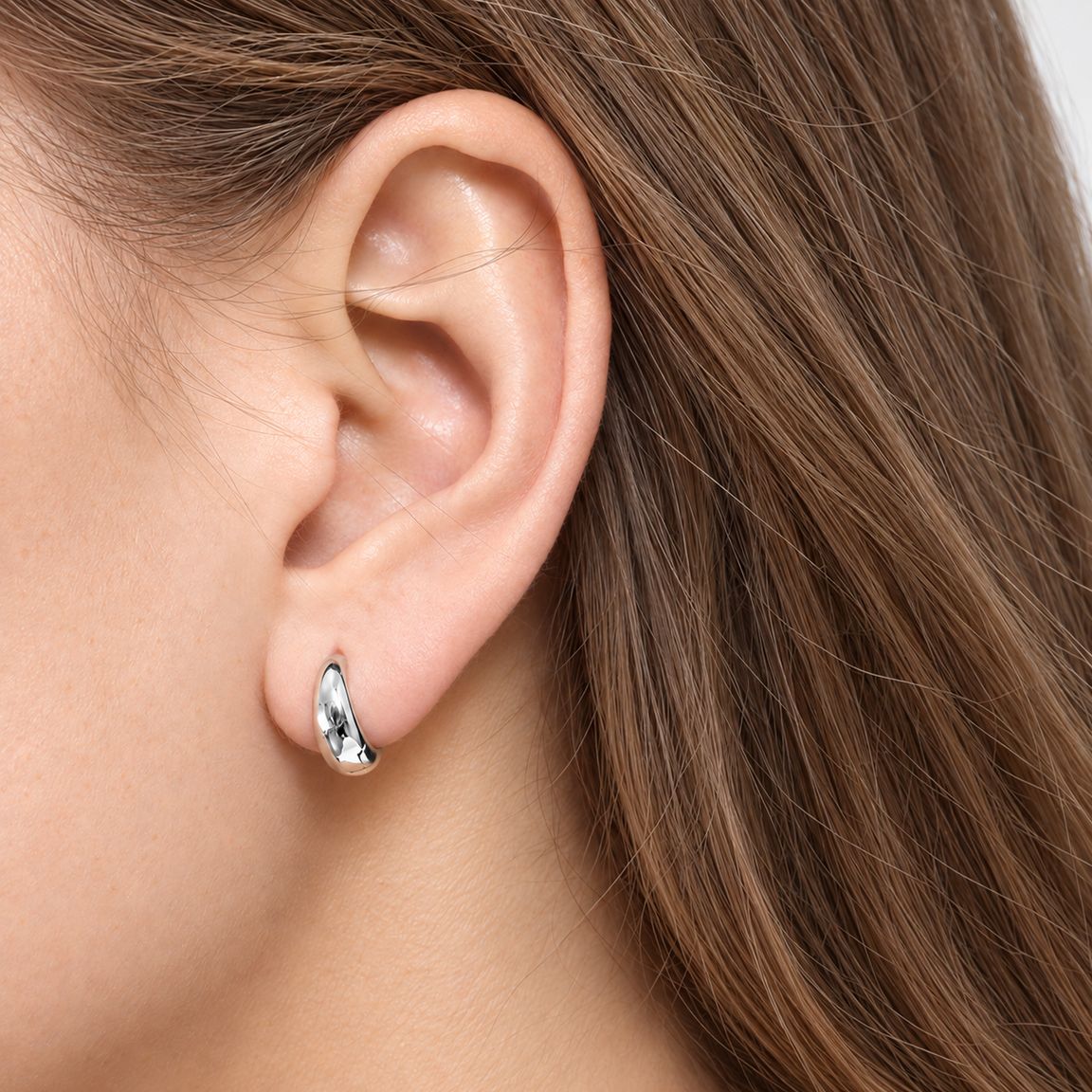 Woman wearing an 8 mm silver teardrop huggie hoop on her earlobe, close side view with loose light-brown hair