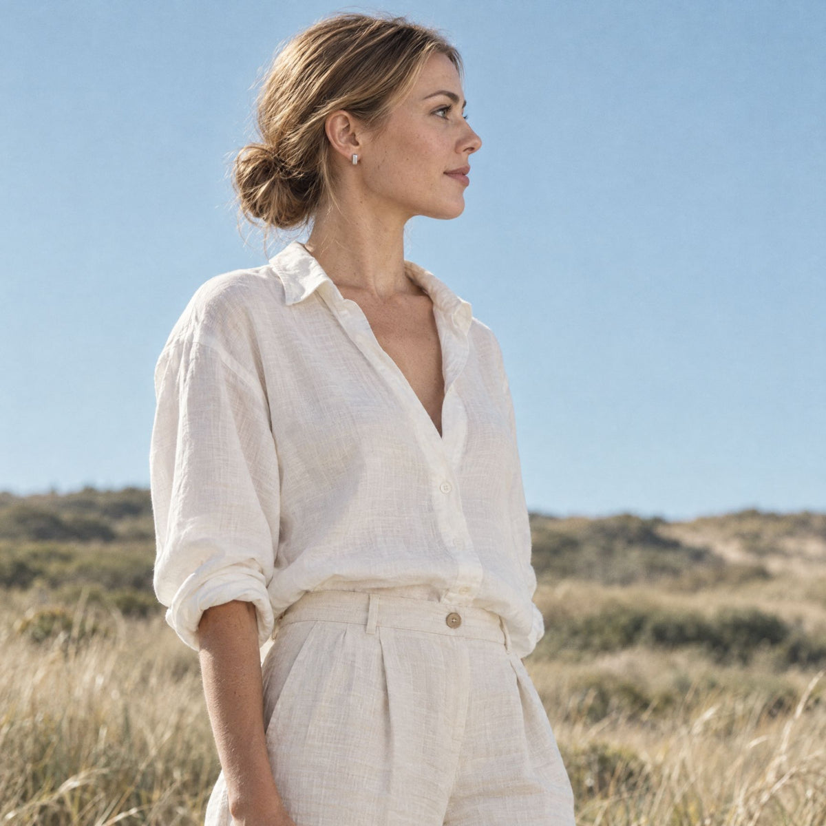 Woman in a cream shirt wearing a small sterling silver huggie earring outdoors in dune grass under a blue sky