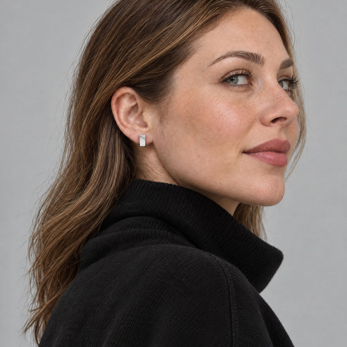 Woman wearing a small sterling silver rectangle huggie earring close to the lobe, side profile on a grey studio ground