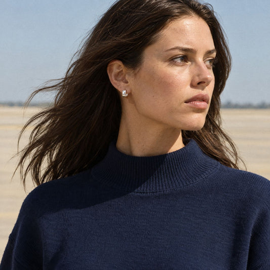 Woman in a navy knit sweater wearing a small square sterling silver huggie earring, outdoors in bright sun