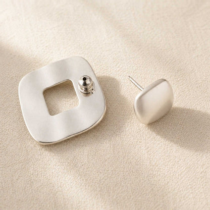 Matte sterling silver stud earrings shown from behind, open-square frame piece and cushion stud on cream linen