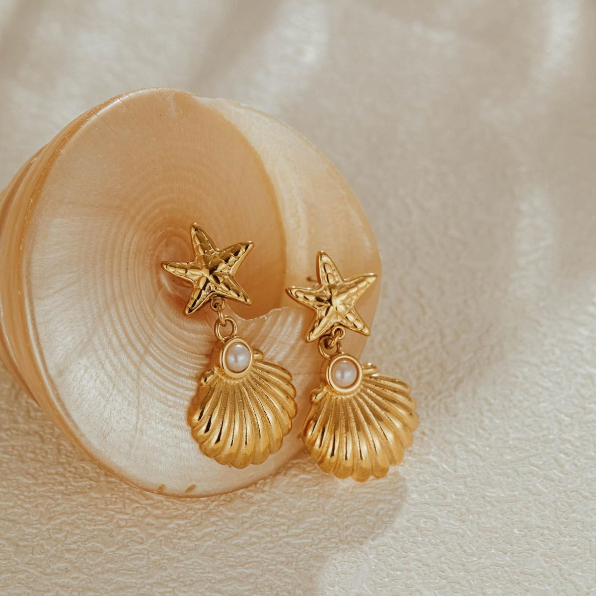 Two gold plated starfish and shell pearl drop earrings resting inside a pale spiral seashell in soft light