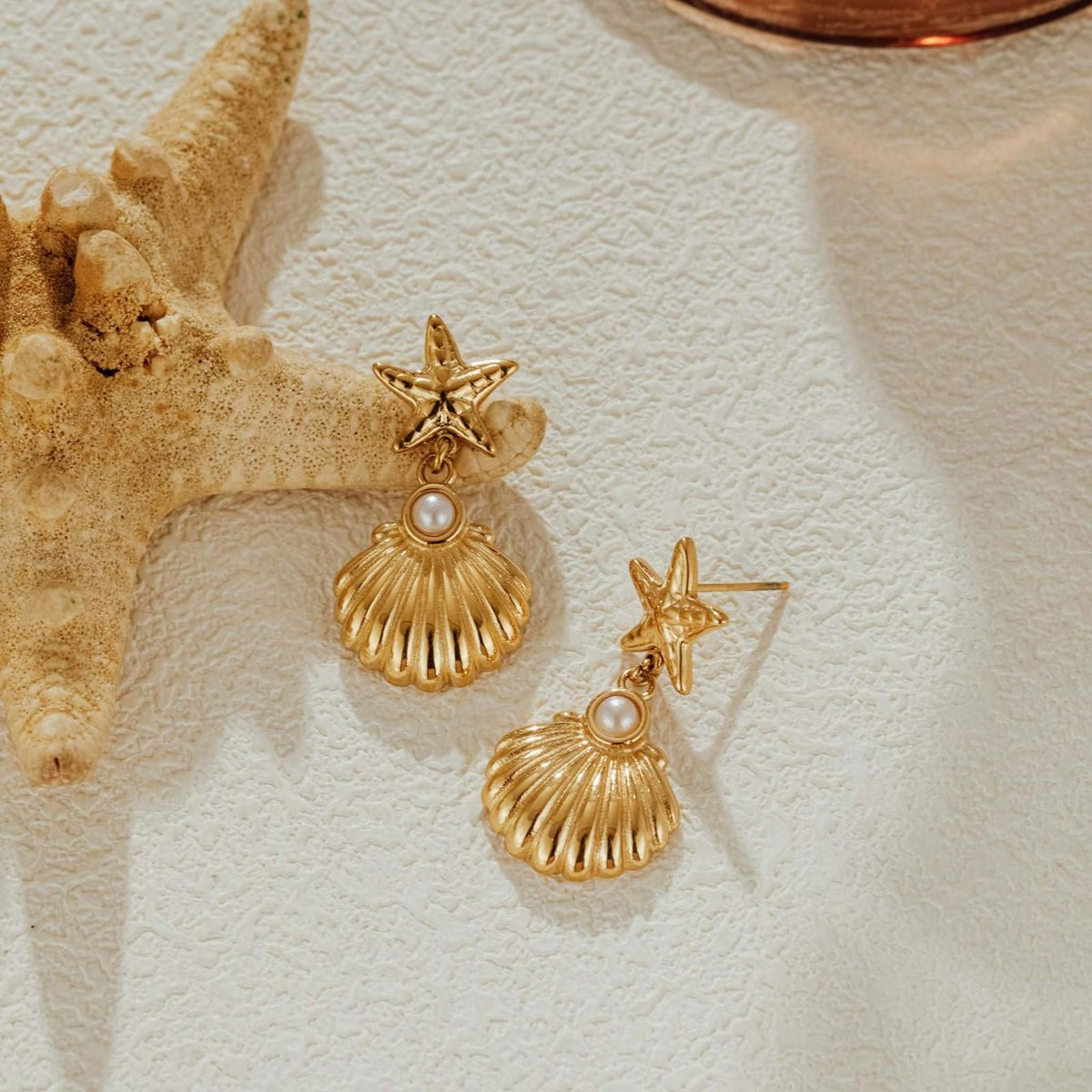 Gold starfish stud seashell drop earrings beside a natural starfish on a textured white surface, one turned