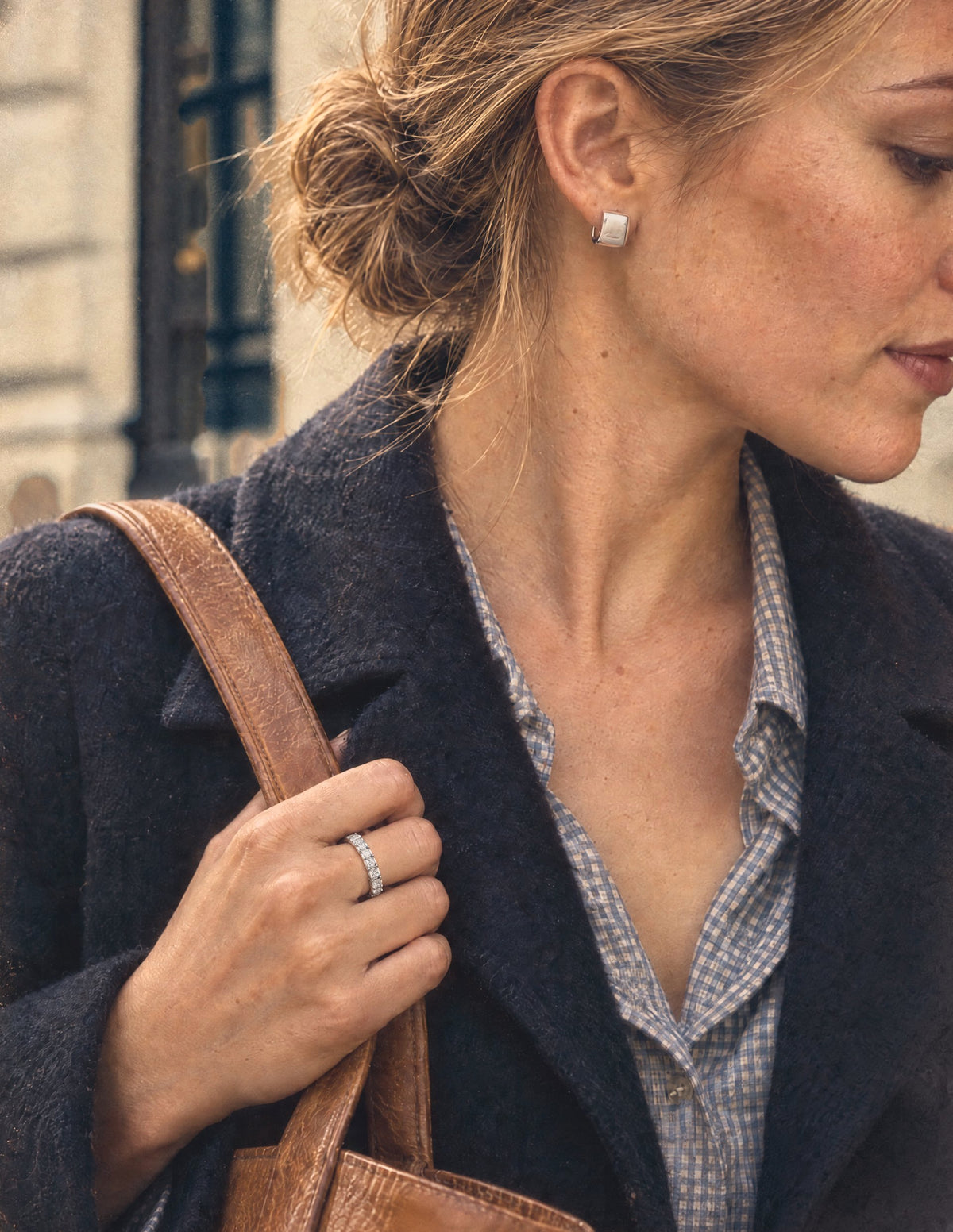 Woman in a navy wool coat on a city street gripping a leather bag strap, wearing a sterling silver eternity ring and square huggie earrings