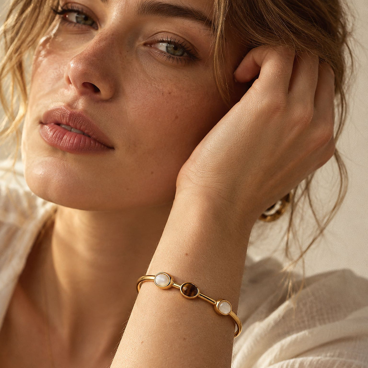 Woman with tousled blonde hair wearing a thin gold tiger eye cuff bracelet, her wrist raised beside her face