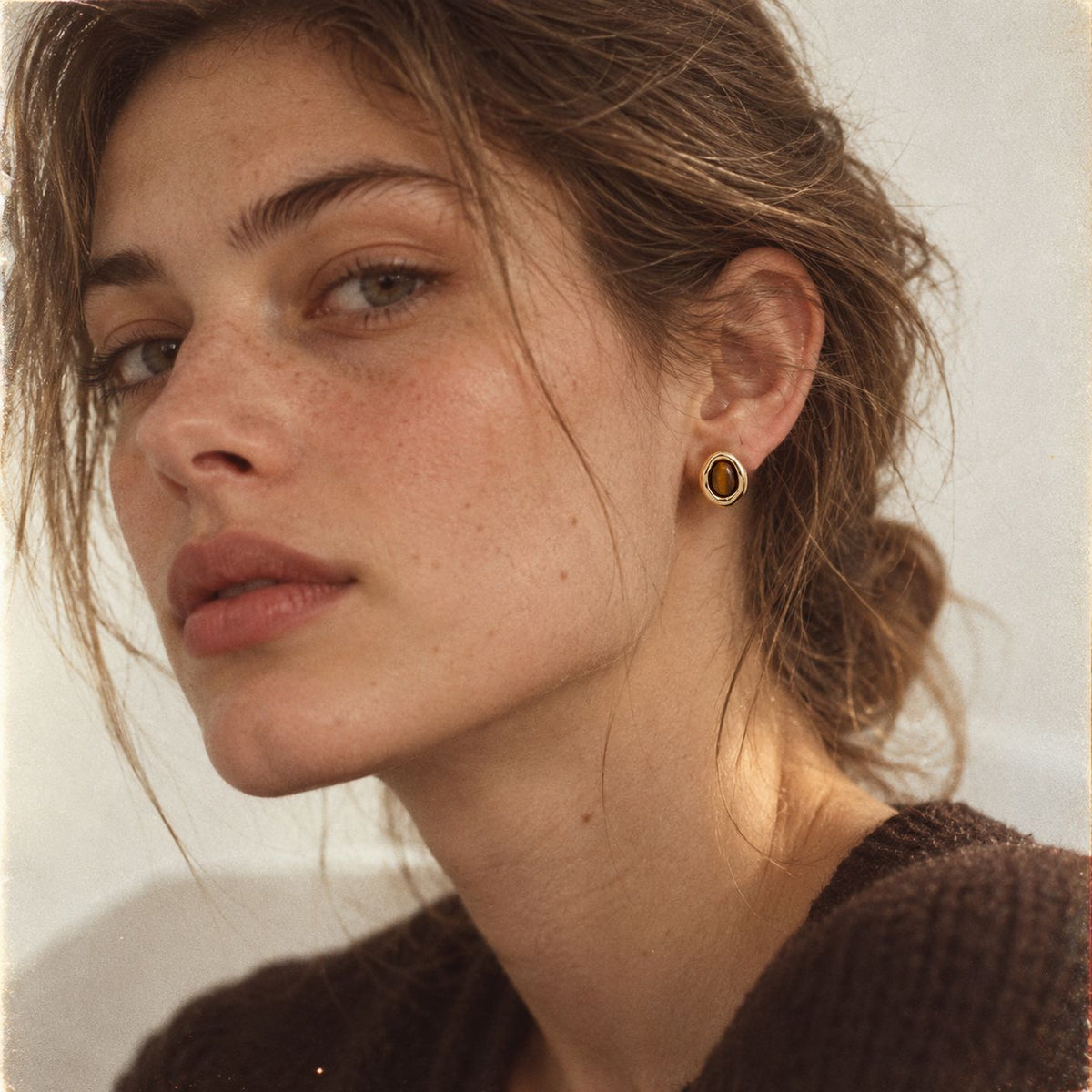 Freckled woman with a messy updo wearing a gold-plated tiger eye stud earring, three-quarter view, brown knit sweater