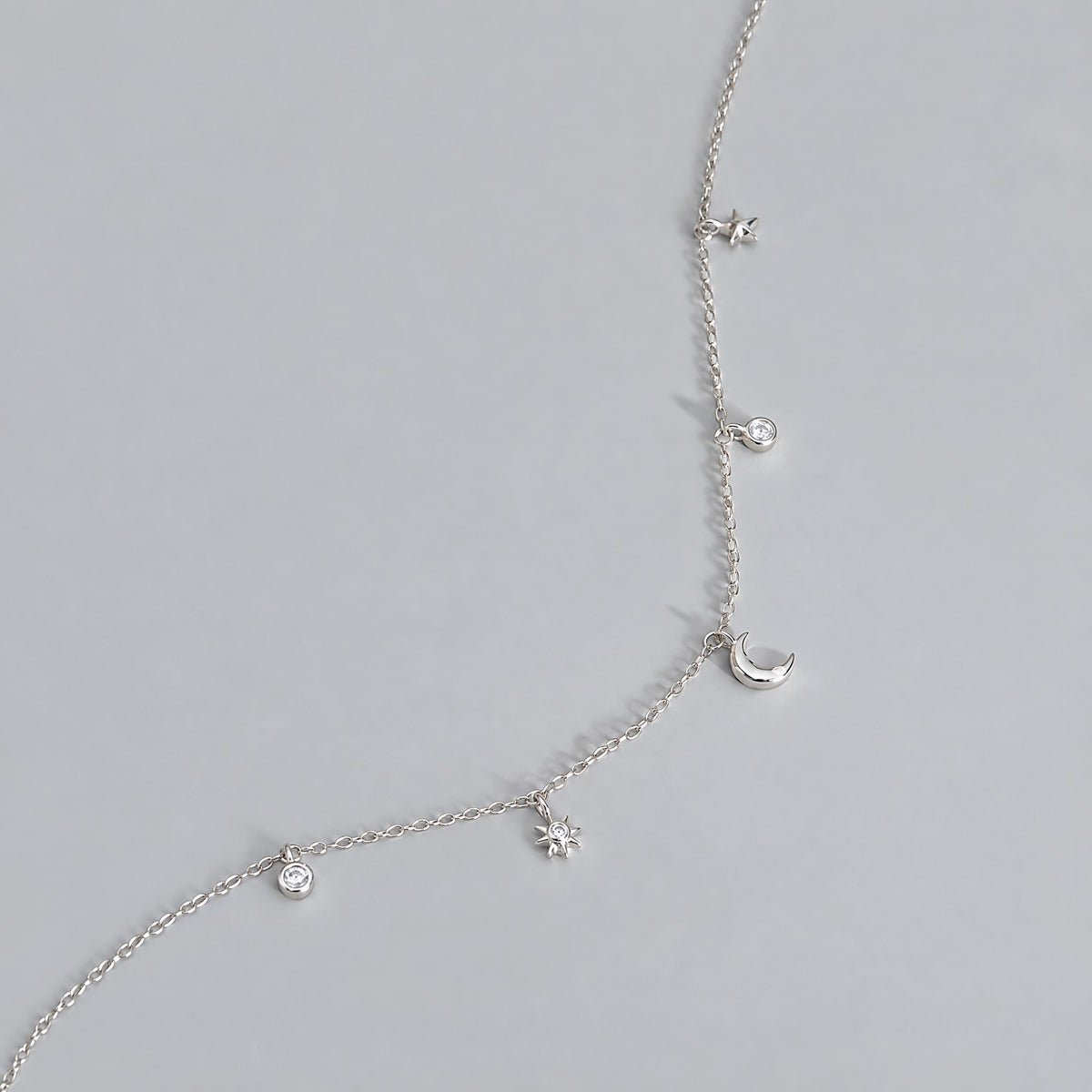 Close up of white gold plated moon and star necklace charms on a dove grey backdrop, crescent moon and starburst