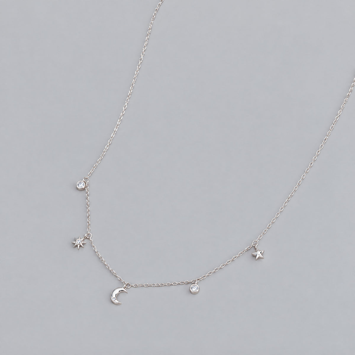 White gold plated moon and star charm necklace laid flat on a dove grey backdrop, crescent and round stone charms