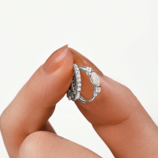 Fingers holding a pair of white gold opal huggie hoop earrings up close, marquise opal and cubic zirconia lined band